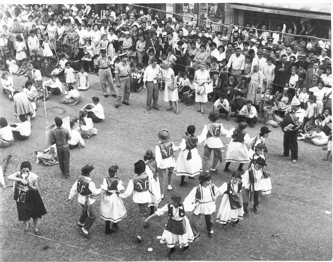 Svartvitt historiskt fotografi av barn i folkdräkter som dansar i en ring på ett torg, omgivna av åskådare.